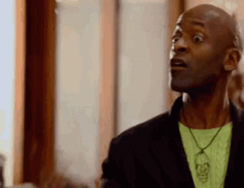 A Man Wearing A Black Jacket And A Green Shirt With A Skull Necklace Is Making A Surprised Face . GIF