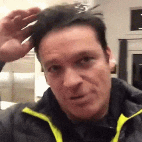 A Man Wearing A Black Jacket With A Yellow Collar Is Holding A Pair Of Scissors To His Head . GIF