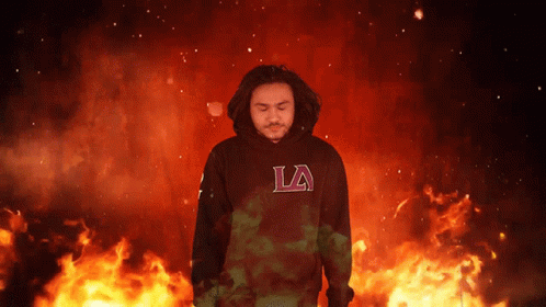 a man wearing a black la hoodie stands in front of flames