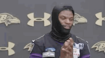 a man wearing a black mask and a purple jersey is standing in front of a wall with ravens logos .