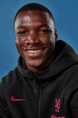A Man Wearing A Black Nike Hoodie Smiles For The Camera Against A Blue Background GIF