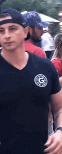 A Man Wearing A Black Shirt And A Baseball Cap Is Standing In A Crowd Of People . GIF