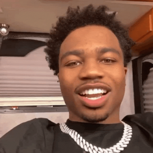 A Man Wearing A Black Shirt And A Silver Chain Is Smiling For The Camera GIF