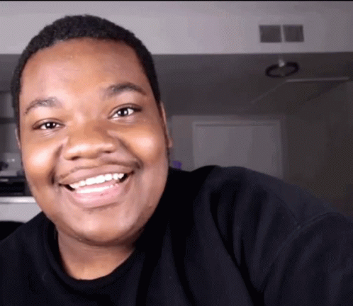 A Man Wearing A Black Shirt Is Smiling And Looking At The Camera GIF