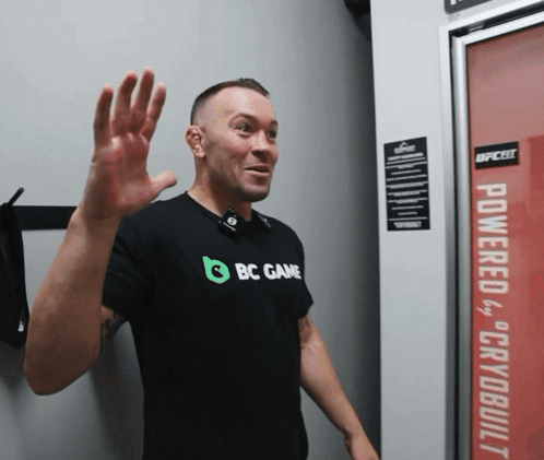 A Man Wearing A Black Shirt That Says Bc Game Waves His Hand GIF