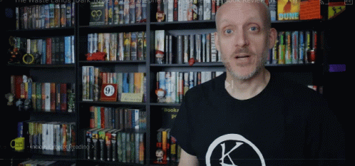 A Man Wearing A Black Shirt With A K On It Stands In Front Of A Bookshelf GIF
