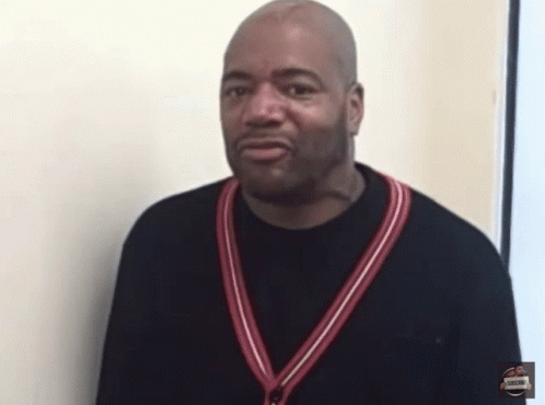 A Man Wearing A Black Shirt With A Red And White Striped Sweater Around His Neck Stands In Front Of A White Wall GIF