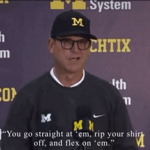 A Man Wearing A Black Shirt With The Letter M On It Is Talking Into A Microphone GIF