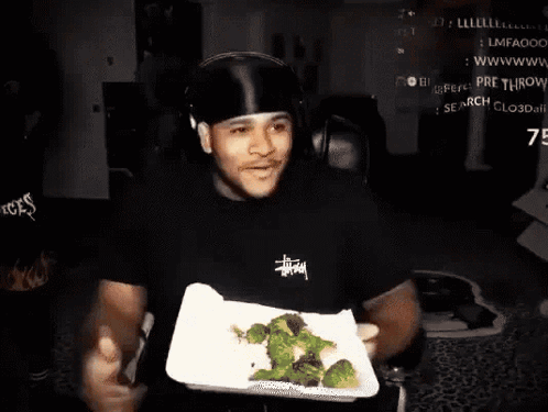 a man wearing a black stussy shirt is holding a plate of broccoli in his hands