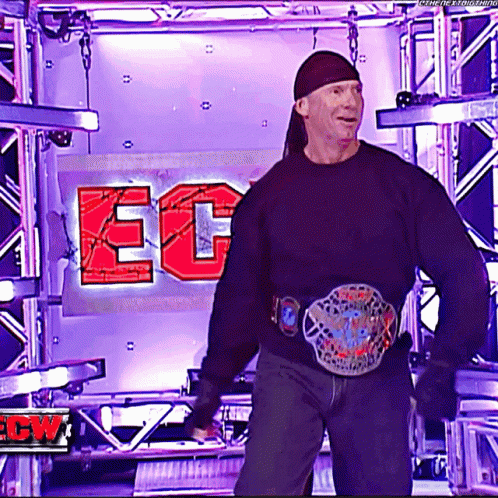 A Man Wearing A Black Sweater And A Belt Is Standing In Front Of A Sign That Says Ec On It GIF
