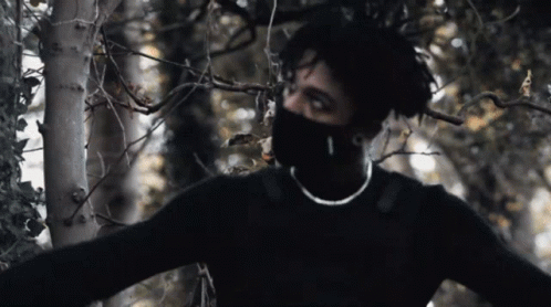 A Man Wearing A Black Sweater And A Mask Is Standing In A Forest GIF