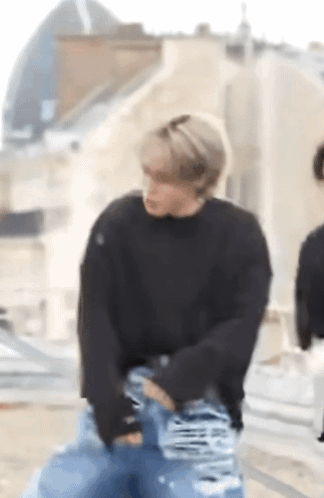 A Man Wearing A Black Sweater And Blue Jeans Is Standing On A Sidewalk . GIF