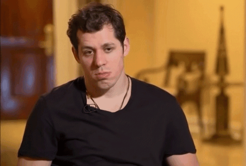A Man Wearing A Black T-Shirt Is Sitting In Front Of A Chair GIF