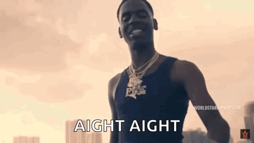 A Man Wearing A Black Tank Top And A Necklace Is Standing In Front Of A Sunset . GIF