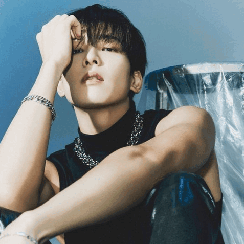 A Man Wearing A Black Tank Top And A Silver Chain Around His Neck Is Sitting Down GIF