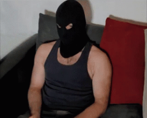 A Man Wearing A Black Tank Top And A Ski Mask Sits On A Couch GIF