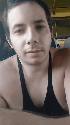 A Man Wearing A Black Tank Top Looks At The Camera With A Yellow Fan In The Background GIF