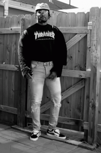 A Man Wearing A Black Thrasher Shirt Stands In Front Of A Wooden Fence GIF