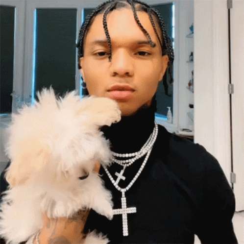 A Man Wearing A Black Turtleneck And A Necklace With A Cross On It Holds A Small White Dog GIF