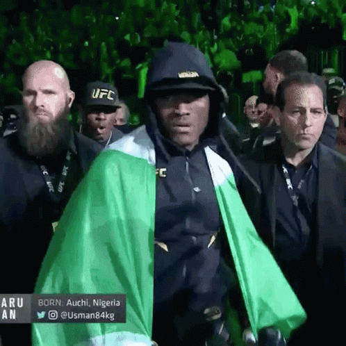 A Man Wearing A Black Ufc Hat And A Green Cape Walks Through A Crowd GIF
