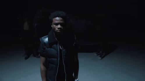 A Man Wearing A Black Vest And Headphones Stands In The Dark GIF