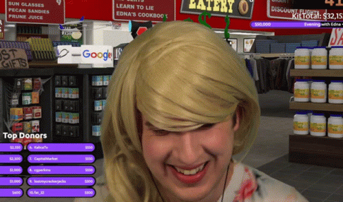 A Man Wearing A Blonde Wig Is Smiling In Front Of A Sign That Says Eatery GIF