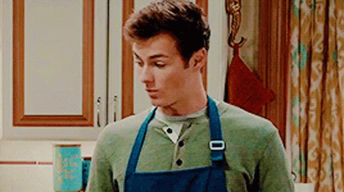 A Man Wearing A Blue Apron Is Standing In A Kitchen . GIF