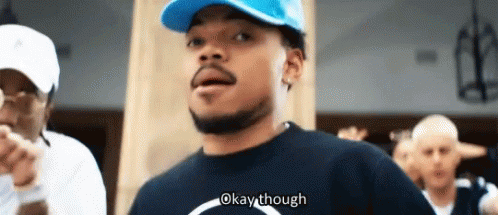 A Man Wearing A Blue Hat And A Black Shirt Is Standing In Front Of A Group Of People And Saying Okay Though . GIF