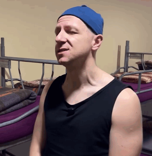 A Man Wearing A Blue Hat And A Black Tank Top Is Standing In Front Of Bunk Beds GIF