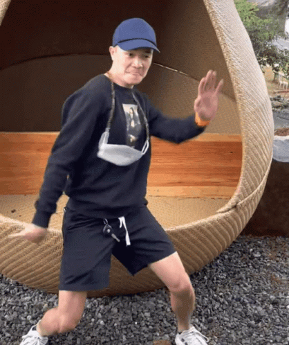 A Man Wearing A Blue Hat And A Face Mask Is Standing In Front Of A Wicker Chair GIF