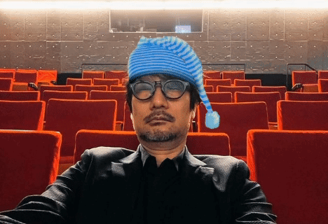 A Man Wearing A Blue Hat And Glasses Sits In A Theater GIF