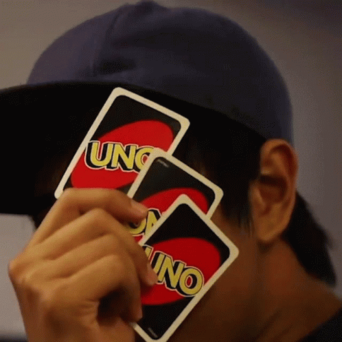 A Man Wearing A Blue Hat Holds Two Uno Cards In Front Of His Face GIF