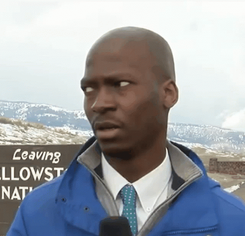 A Man Wearing A Blue Jacket And Tie Stands In Front Of A Sign That Says Leaving Yellowstone National Park GIF