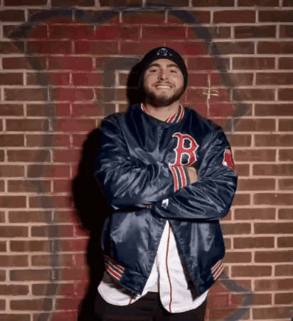 A Man Wearing A Blue Jacket With The Letter B On It Stands In Front Of A Brick Wall GIF