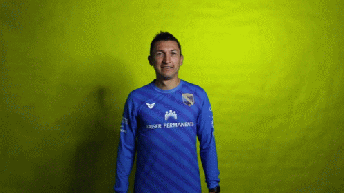 A Man Wearing A Blue Kaiser Permanent Jersey Stands In Front Of A Yellow Background GIF