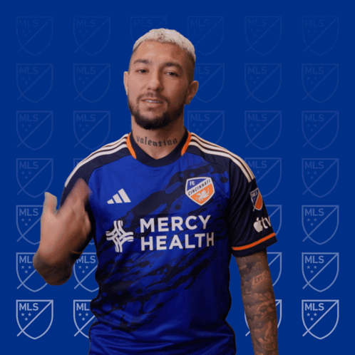A Man Wearing A Blue Mercy Health Jersey Stands In Front Of A Blue Background With Mls Logos GIF