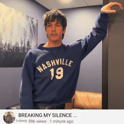 a man wearing a blue nashville 19 sweatshirt leaning against a wall