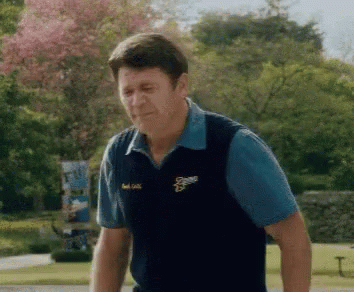 A Man Wearing A Blue Shirt And A Black Vest With The Letter S On It Is Standing In A Park . GIF