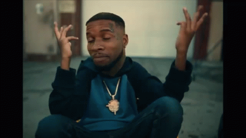 A Man Wearing A Blue Shirt And A Gold Necklace Is Sitting On The Ground With His Hands Up GIF
