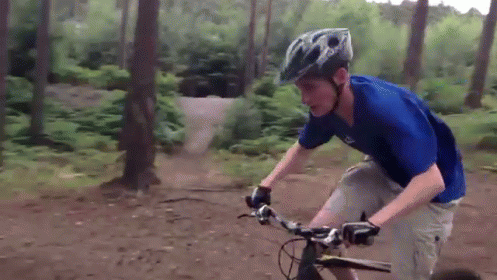 A Man Wearing A Blue Shirt And A Helmet Is Riding A Bike On A Dirt Road GIF