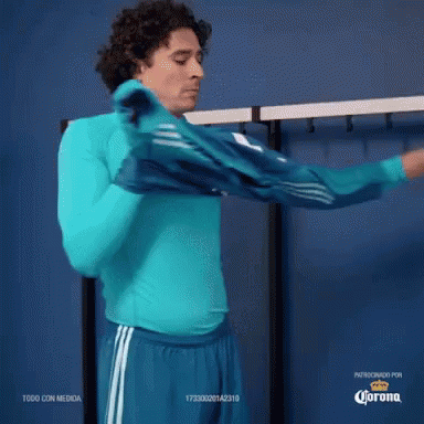 A Man Wearing A Blue Shirt And Blue Shorts Is Standing In A Locker Room With Corona Written On The Bottom GIF