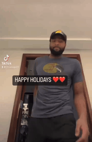 A Man Wearing A Blue Shirt That Says Happy Holidays Is Standing In Front Of A Door GIF