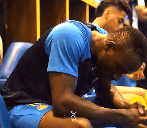 A Man Wearing A Blue Shirt With The Letter X On It Sits On A Chair GIF