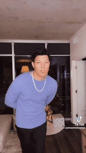 A Man Wearing A Blue Sweater And A Gold Chain Is Standing In A Living Room .. GIF