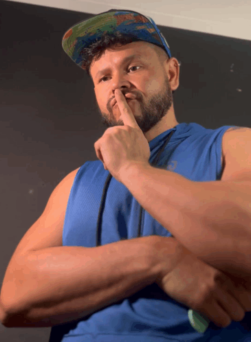 A Man Wearing A Blue Tank Top And A Hat Holds His Finger To His Nose GIF