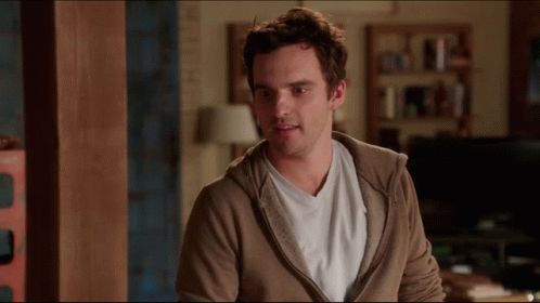 A Man Wearing A Brown Hoodie And A White Shirt Stands In A Living Room GIF