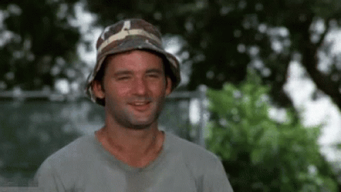 A Man Wearing A Bucket Hat And A Gray Shirt Is Smiling And Looking At The Camera . GIF