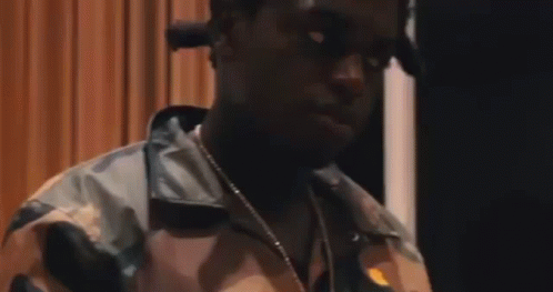A Man Wearing A Camouflage Jacket And A Necklace Is Standing In Front Of A Curtain . GIF