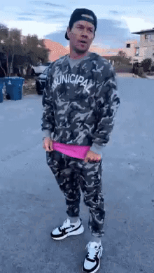 A Man Wearing A Camouflage Sweater And Pants Is Standing On A Street . GIF