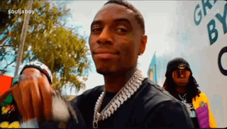 A Man Wearing A Chain Around His Neck Is Smiling While Standing In Front Of A Wall . GIF
